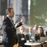 Un señor esta hablando delante de las personas, dictando una conferencia, desarrollando un evento de emprendedores y periodistas.