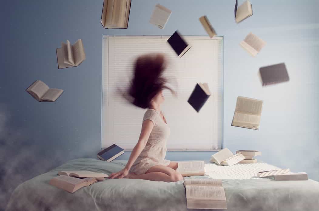 Una mujer esta sentada encima de su cama con su cabello alborotado y un montón de libros al aire, aprovecha la feria del libro en bogotá, y con ella la gran pregunta para qué ir a la feria y visitarla