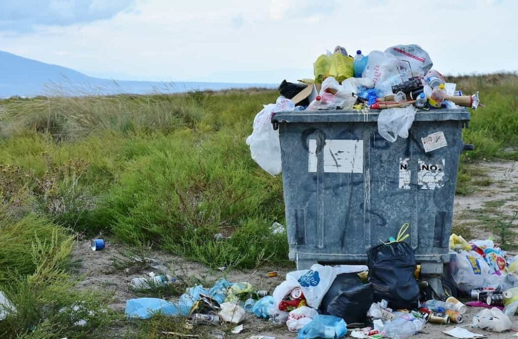 Un bote de basura en medios de un prado verde, toda la basura esta en el piso, cuidemos la madre tierra, nuestro hogar.