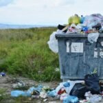 Un bote de basura en medios de un prado verde, toda la basura esta en el piso, cuidemos la madre tierra, nuestro hogar.