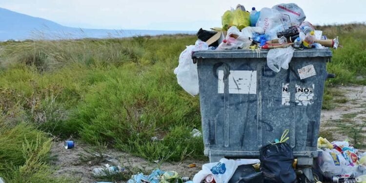 Un bote de basura en medios de un prado verde, toda la basura esta en el piso, cuidemos la madre tierra, nuestro hogar.