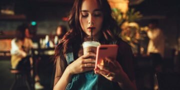 Chica en una tienda, mirando su celular y tomando cafe.