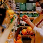 Una mujer con tapabocas haciendo mercado y agarrando una fruta.