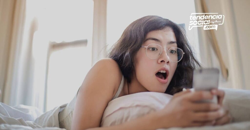 Mujer acostada en la cama sorprendida mirando su celular