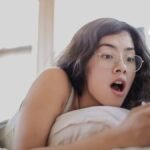 Mujer acostada en la cama sorprendida mirando su celular